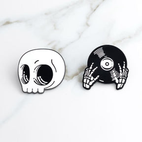 The white skull and black record-hands pins arranged separately on pale marble, showing their contrasting shapes.