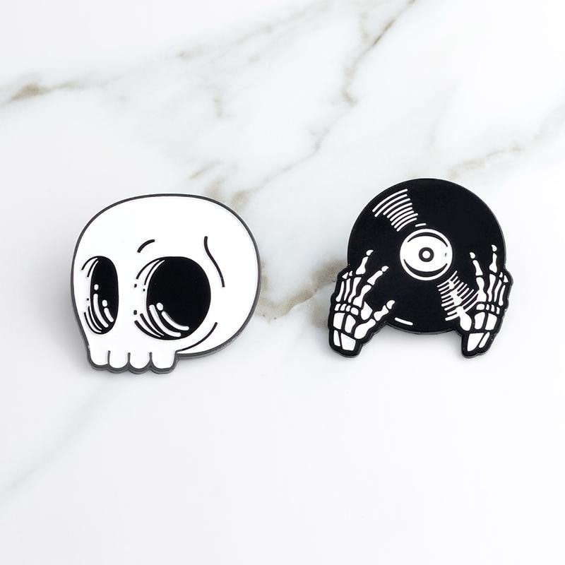 The white skull and black record-hands pins arranged separately on pale marble, showing their contrasting shapes.