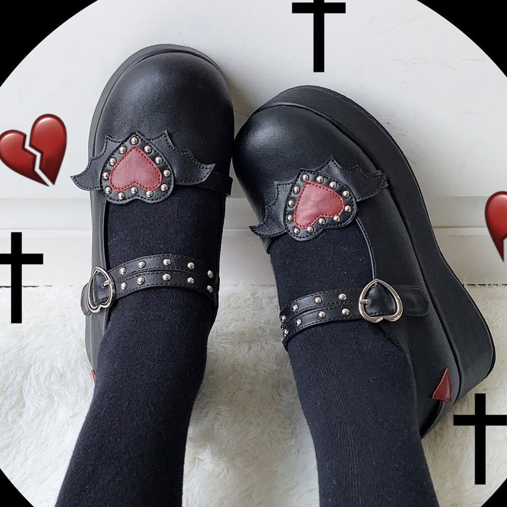 Black winged-heart Mary Janes worn with black socks, viewed from above against a graphic background with crosses and red hearts.