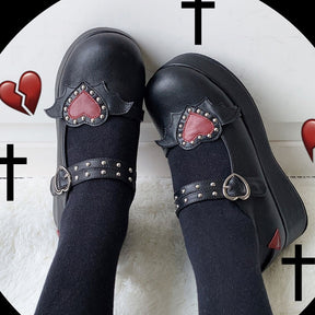 Black winged-heart Mary Janes worn with black socks, viewed from above against a graphic background with crosses and red hearts.