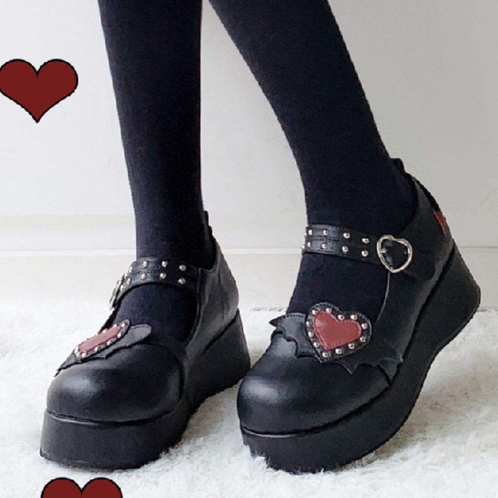 Black winged-heart platform Mary Janes worn with black tights, shown from the front with the red studded hearts and buckled straps visible.