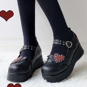 Black winged-heart platform Mary Janes worn with black tights, shown from the front with the red studded hearts and buckled straps visible.