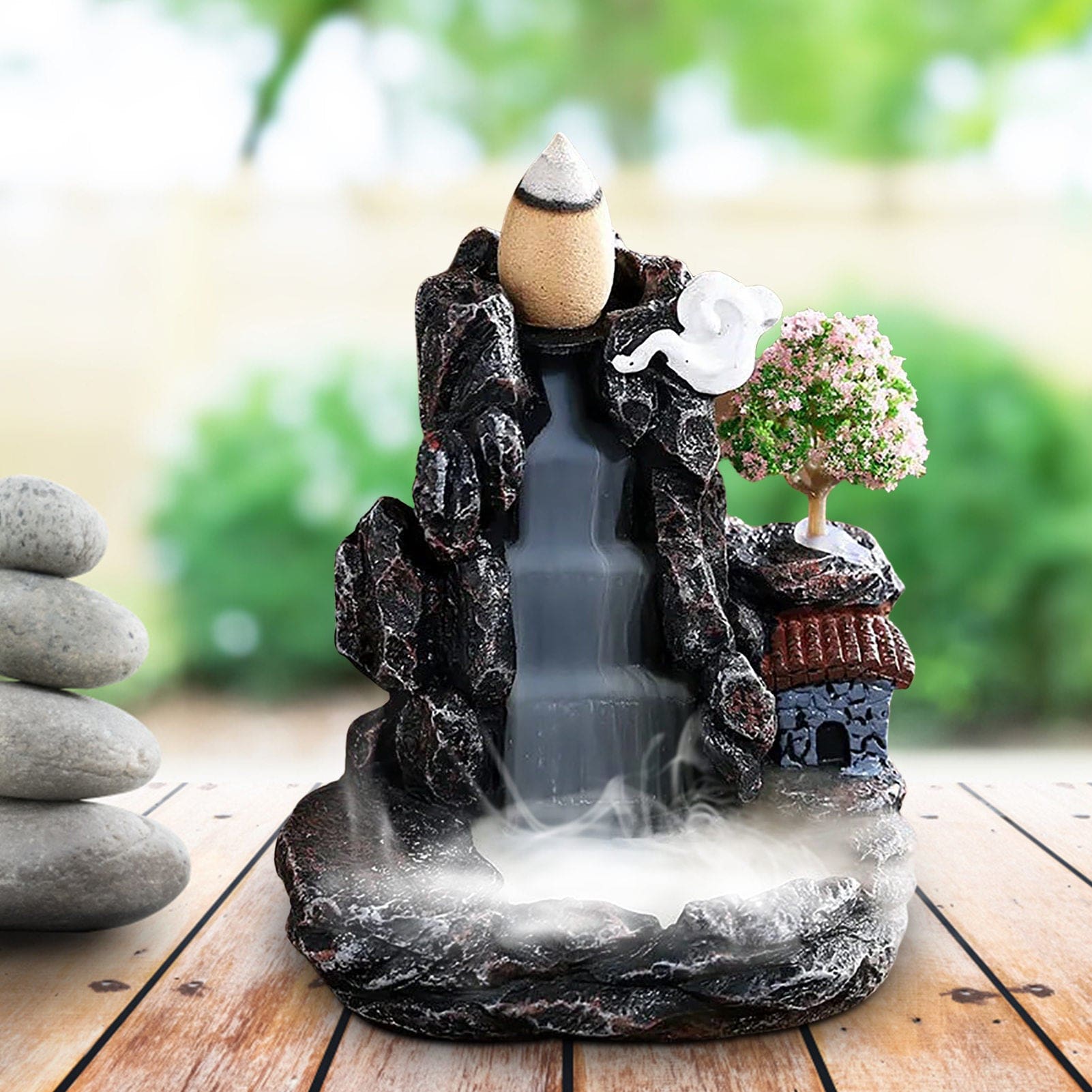 Village backflow burner displayed beside smooth stacked stones and green plants, with pale smoke flowing down the dark central channel.