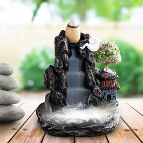 Village backflow burner displayed beside smooth stacked stones and green plants, with pale smoke flowing down the dark central channel.