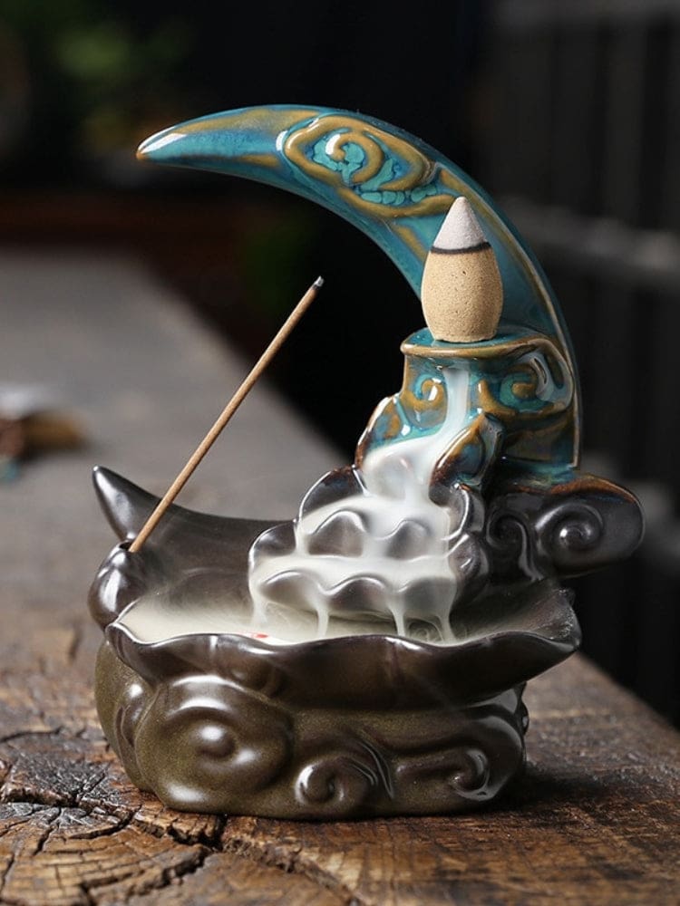 Blue crescent moon backflow incense burner on a rustic wooden surface, with a cone on the raised platform and white smoke spilling down the sculpted tiers into a dark curved basin. An incense stick rests beside the smoke path.