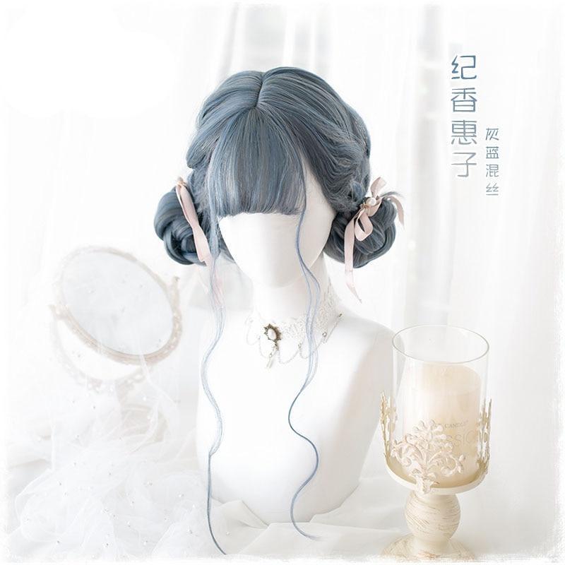 Blue-gray wig styled in two low rolled buns with a full fringe and loose face-framing strands. Supplier lettering appears beside the mannequin in the pale decorative setting.