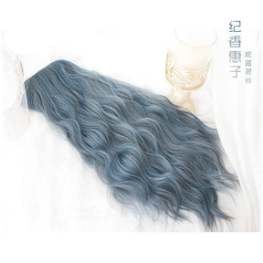 Blue-gray wig laid across a pale surface with the crown to the left and long wavy strands spread toward the right, showing the blended color and wave texture.