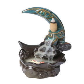 Blue crescent moon backflow burner isolated against white, showing gold-toned scroll accents, a small fish detail inside the dark basin and smoke flowing from the raised cone.