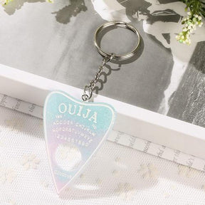Pastel planchette keychain with pale blue and pink tones, white spirit-board lettering and fine sparkle, resting against an open book on white lace.