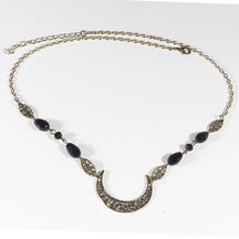 Bronze-tone ornate crescent headdress with large black teardrop beads and small dark accents, arranged in an oval on a pale background with the clasp and chain visible.