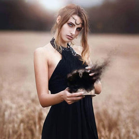 Fuller view of a model in a black dress and crescent forehead chain in dry grass, holding a bowl as dark smoke rises above it.