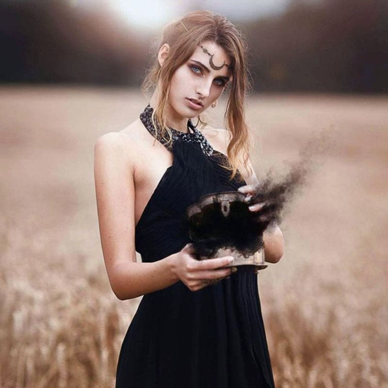 Fuller view of a model in a black dress and crescent forehead chain in dry grass, holding a bowl as dark smoke rises above it.