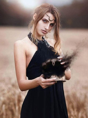 A model in a black halter dress wears a crescent chain headdress in a grassy field while holding a dark smoking bowl.