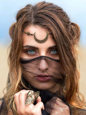Close portrait of a model wearing an ornate bronze-tone crescent headdress at the forehead, with black mesh held across the lower face and an ornate ring beside it.