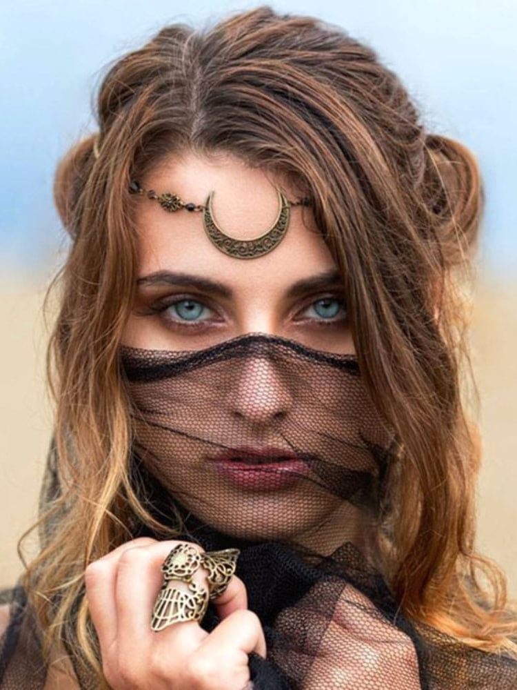 Close portrait of a model wearing an ornate bronze-tone crescent headdress at the forehead, with black mesh held across the lower face and an ornate ring beside it.