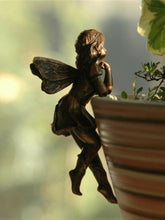 Bronze-tone fairy ornament leaning against a planter rim, with curled hair, patterned wings and bent legs hanging outside the pot.