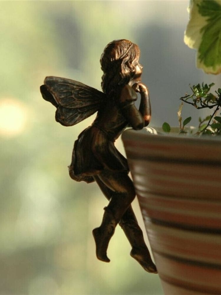 Bronze-tone fairy ornament leaning against a planter rim, with curled hair, patterned wings and bent legs hanging outside the pot.