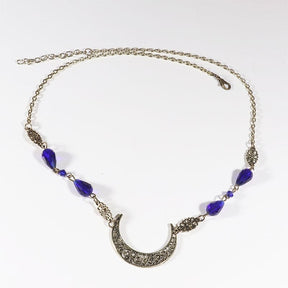 Bronze-tone ornate crescent headdress with large purple teardrop beads, arranged in an oval on a pale background with the clasp and chain visible.