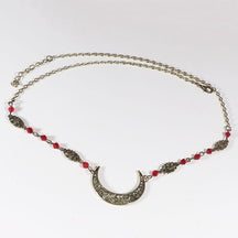 Bronze-tone ornate crescent head chain with small red faceted beads, arranged in an oval on a pale background with the clasp and chain visible.