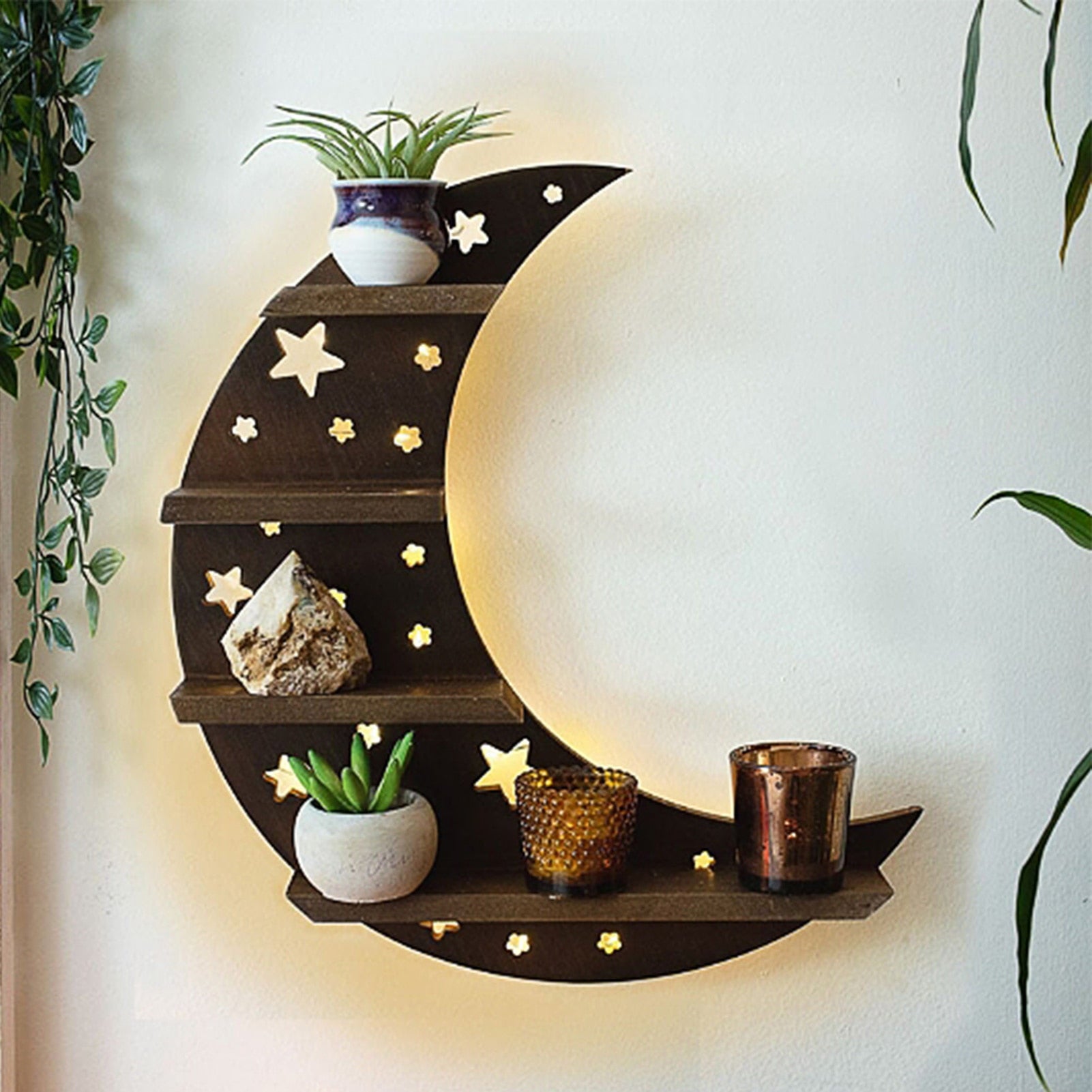 Brown star-cutout crescent shelf mounted on a pale wall beside hanging greenery, with small plants and decorative objects on its four ledges.