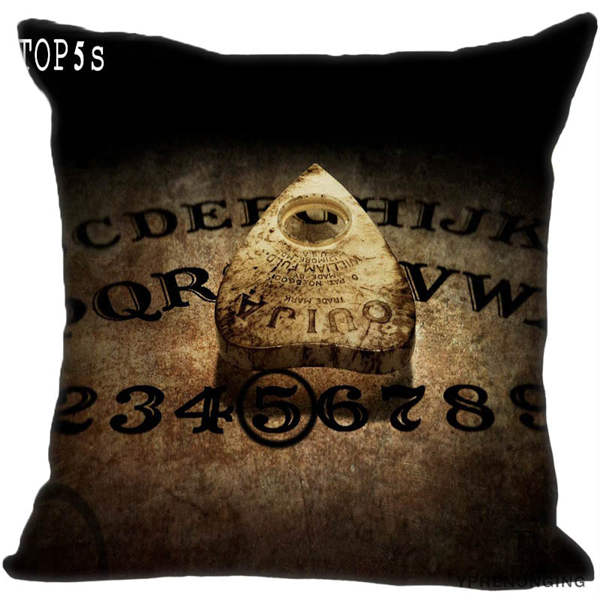 Brown and black square pillow cover printed with a close-up spirit board, alphabet, numbers and a wooden-looking planchette in shadow.