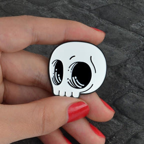 White cartoon skull pin held between fingers against gray fabric, with large black eye sockets and small curved highlights.