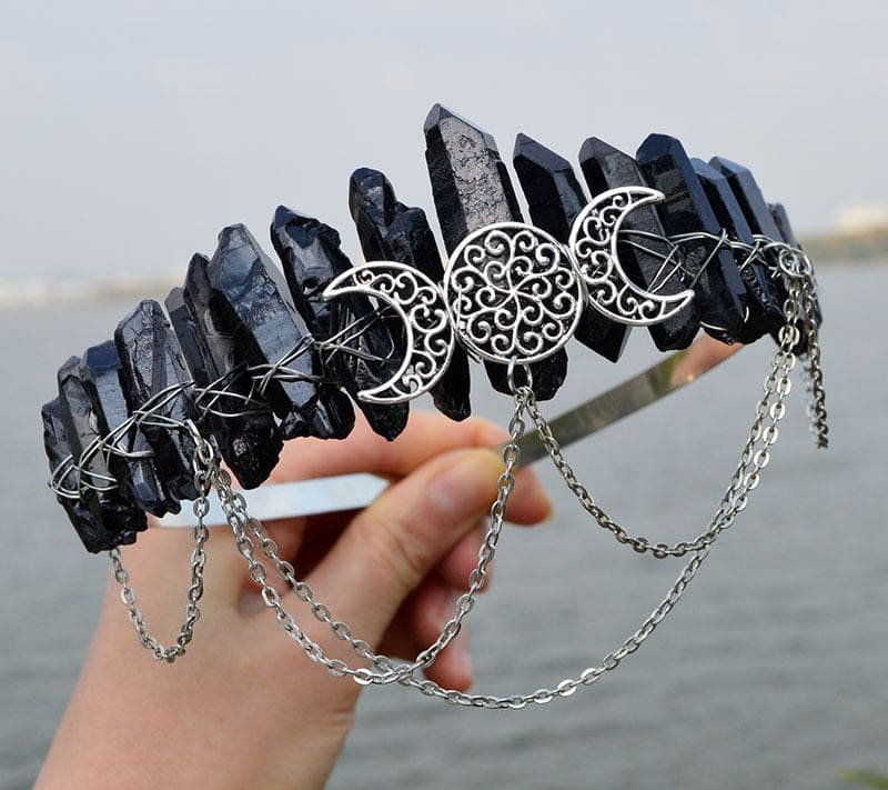 Black crystal crown held beside water, with three filigree moons above draped chains.