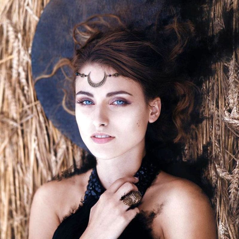 A reclining model wears a crescent chain headdress centred on the forehead, with dark hair spread over a straw-textured background and a black lace neckline below.