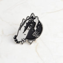 Crystal ball enamel pin on pale marble, showing an illustrated hand over a black orb with tiny celestial marks and silver-tone outlines.