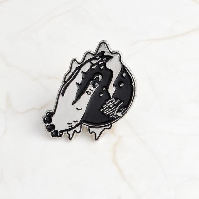 Crystal ball enamel pin on pale marble, showing an illustrated hand over a black orb with tiny celestial marks and silver-tone outlines.