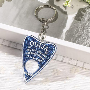 Dark blue glitter planchette resin keychain with white spirit-board lettering and a round cutout, photographed beside an open book on white lace.