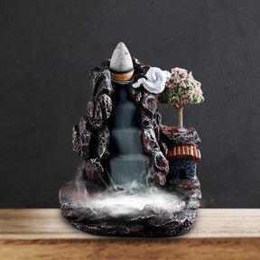 Front view of the dark mountain-village burner on a light wood surface against a black background, with its roof, blossom tree and stepped smoke path visible.