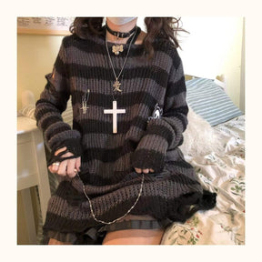 Seated bedroom view of a grey and black long striped sweater, with the sleeves covering part of the hands and a chain lying across the lap.