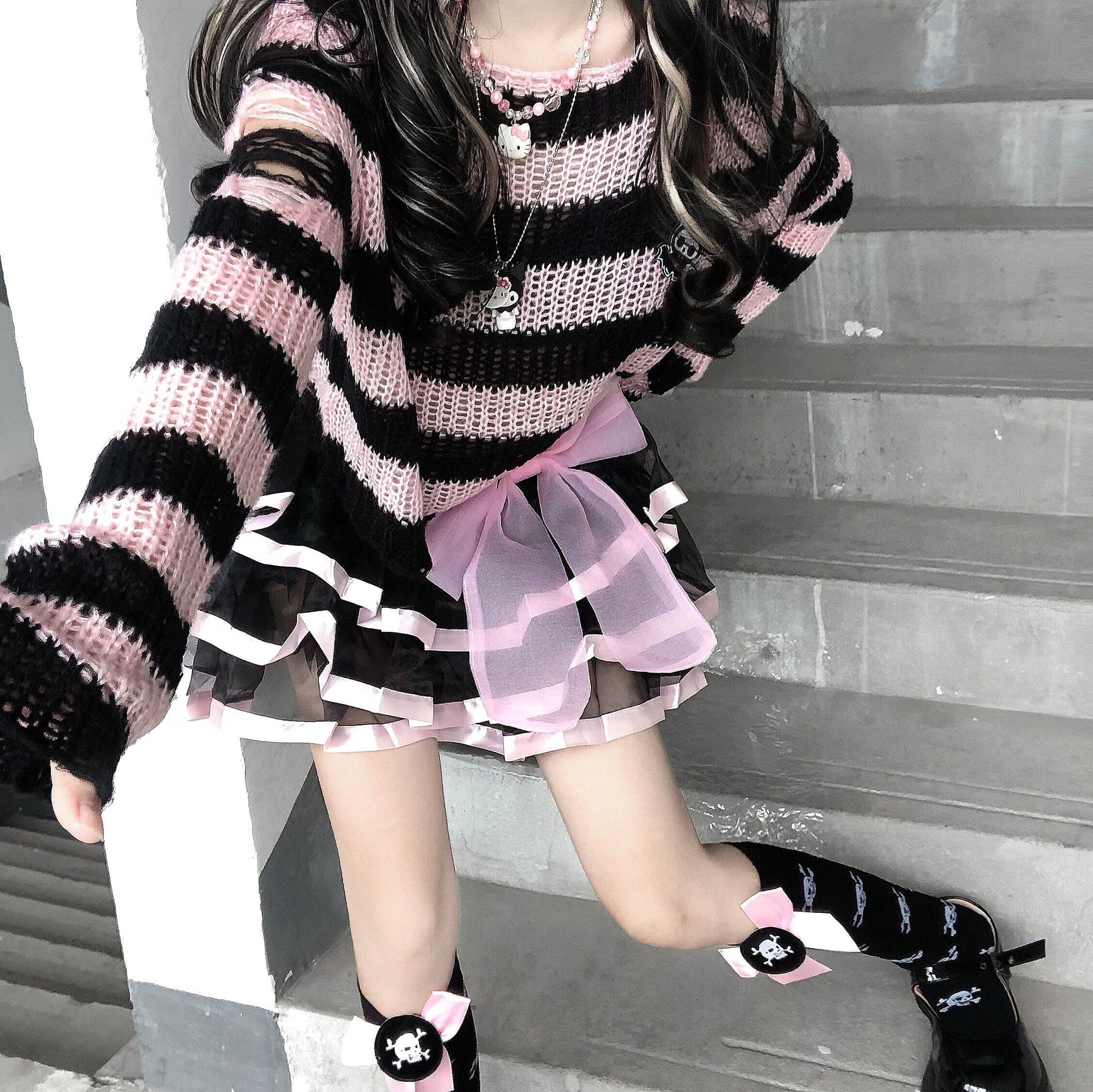 Pink and black cropped sweater seen from above on a staircase, styled with a pink-trimmed skirt, black socks and shoes. The model extends an arm toward the camera.