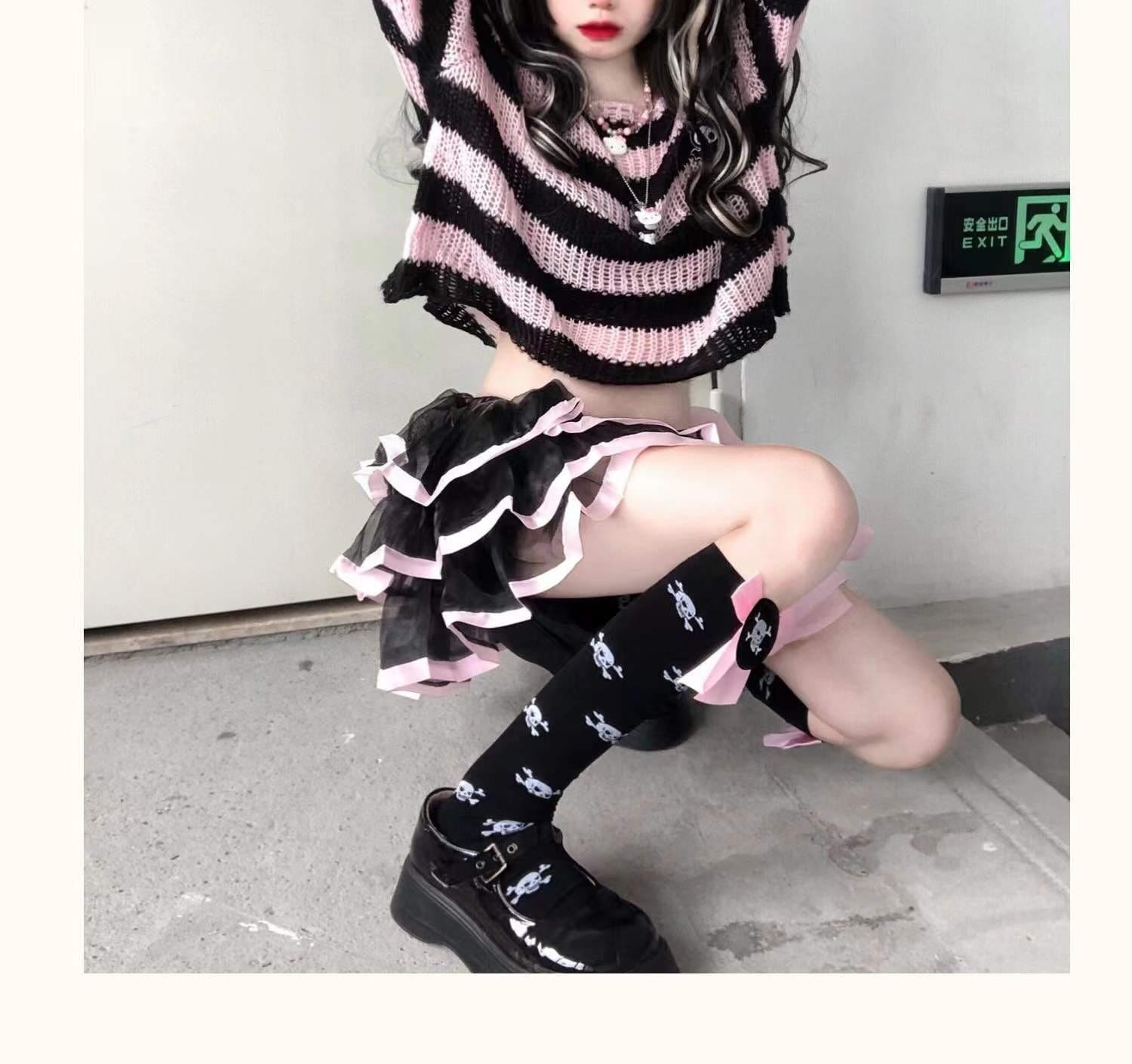 Seated staircase view of a pink and black cropped sweater with a ruffled skirt and black knee socks. One arm is raised above the wide neckline.