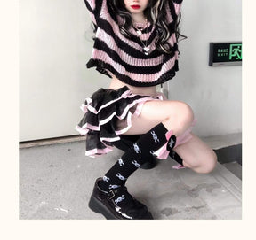 Seated staircase view of a pink and black cropped sweater with a ruffled skirt and black knee socks. One arm is raised above the wide neckline.