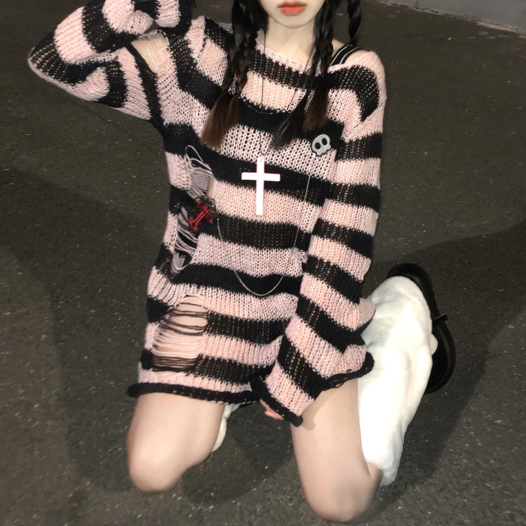Long pink and black striped sweater worn on a staircase, with laddered openings near the lower side and long sleeves. A skull pin and necklace are styled on the front.