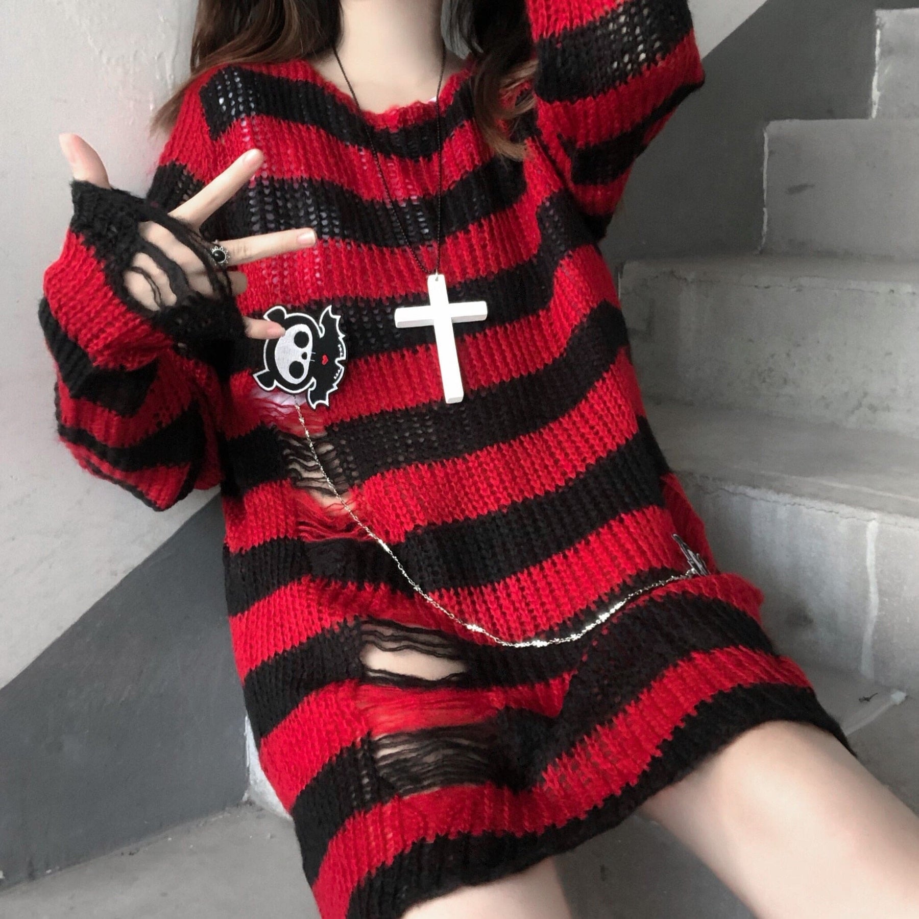 Long red and black striped distressed sweater worn on a staircase, with open laddered sections across the lower front. A cross necklace and small pin are styled over the knit.
