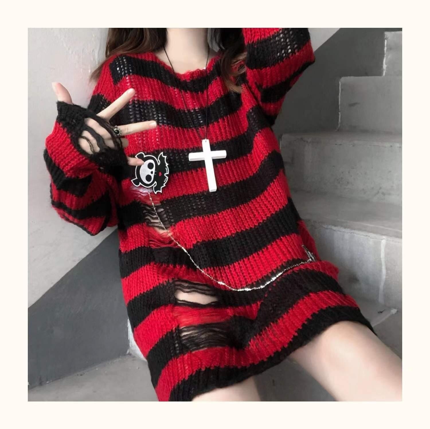 Closer seated view of a red and black long distressed sweater, with laddered openings across the lower front and sleeve cuff. A cross necklace is layered over the stripes.