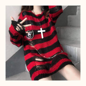 Closer seated view of a red and black long distressed sweater, with laddered openings across the lower front and sleeve cuff. A cross necklace is layered over the stripes.
