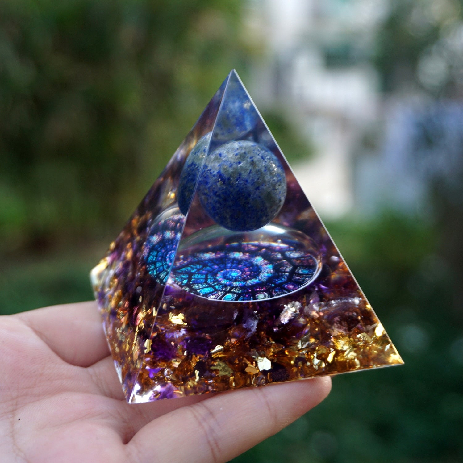 Fibonacci-inspired lapis lazuli orgone pyramid with a blue sphere, spiral inlay and purple amethyst layers. Angled palm view showing the depth of the purple and gold-tone layers.