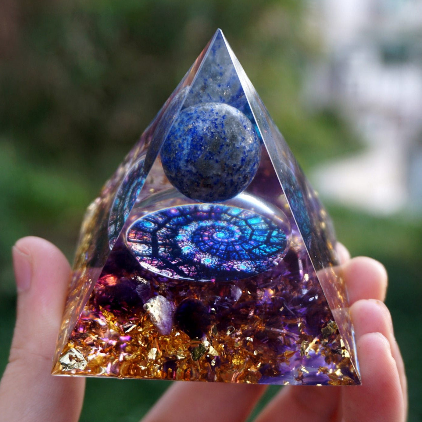 Fibonacci-inspired lapis lazuli orgone pyramid with a blue sphere, spiral inlay and purple amethyst layers. Close front view held outdoors with gold-tone flecks visible below the spiral.