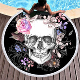 Black round floral skull rug with white fringe spread beside a pool beneath a seated woman. Pink, purple and blue flowers surround the pale skull.