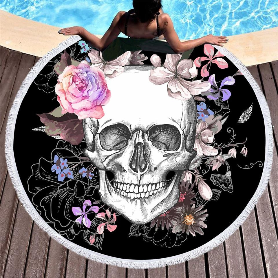 Black round floral skull rug with white fringe spread beside a pool beneath a seated woman. Pink, purple and blue flowers surround the pale skull.