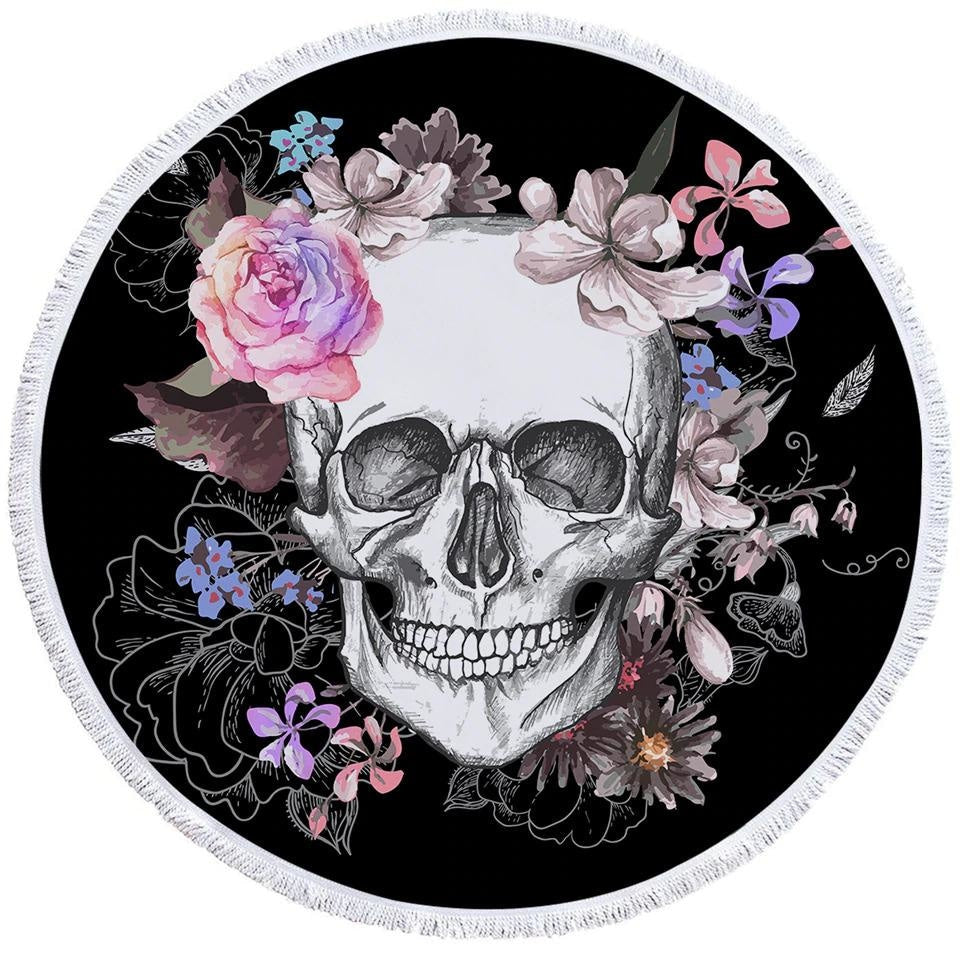 Round black rug shown flat with a large pale skull, pink and purple flowers, green leaves and white fringe around the edge.