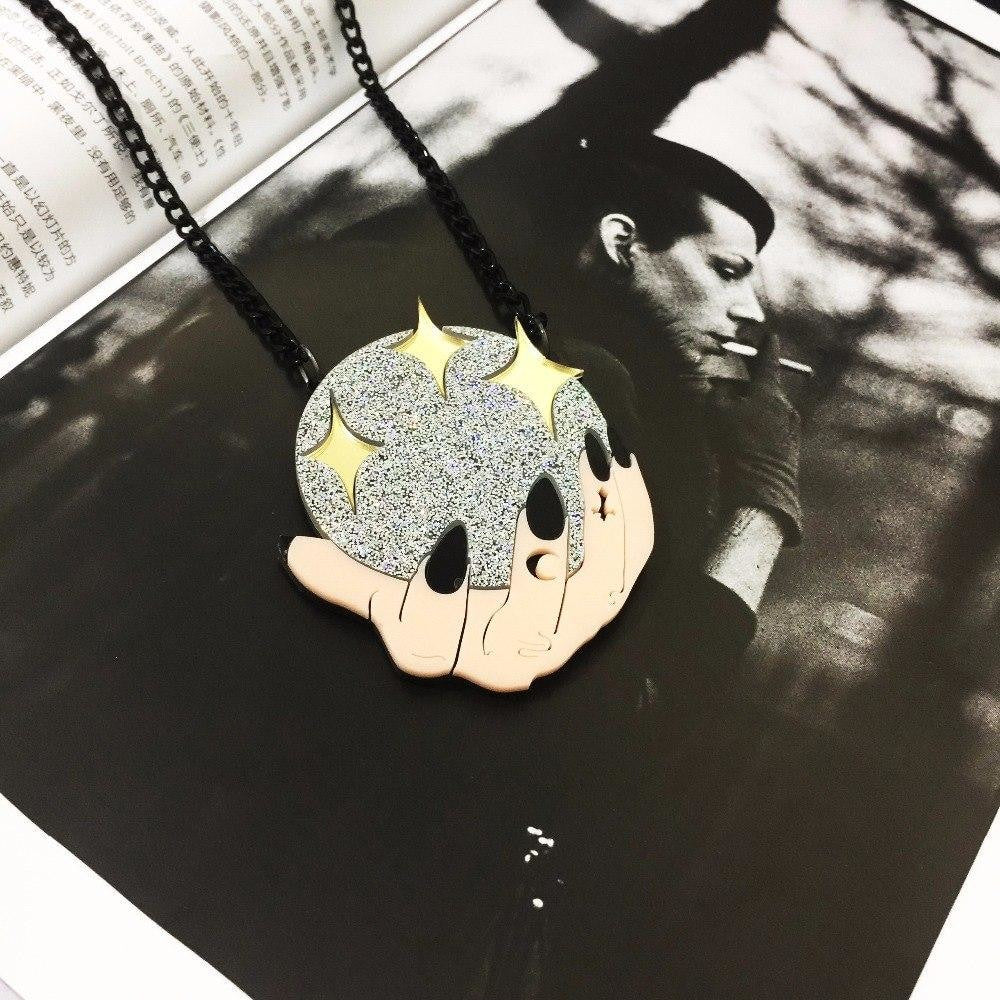 Crystal-ball and hands pendant laid on a black-and-white printed page, with the black chain arranged around the glittering acrylic design.