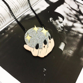 Crystal-ball and hands pendant laid on a black-and-white printed page, with the black chain arranged around the glittering acrylic design.