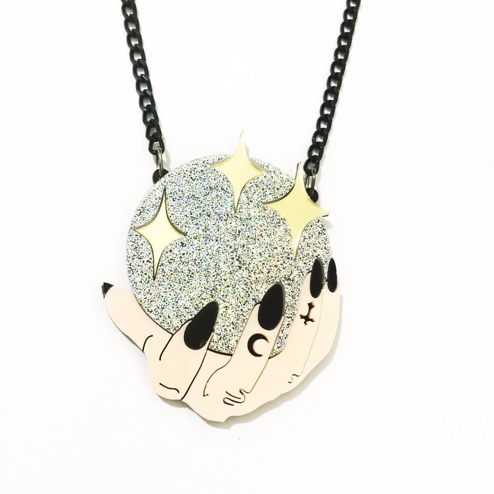 Acrylic pendant necklace depicting pale hands with black nails around a silver glitter crystal ball dotted with gold stars, suspended from a black chain on white.