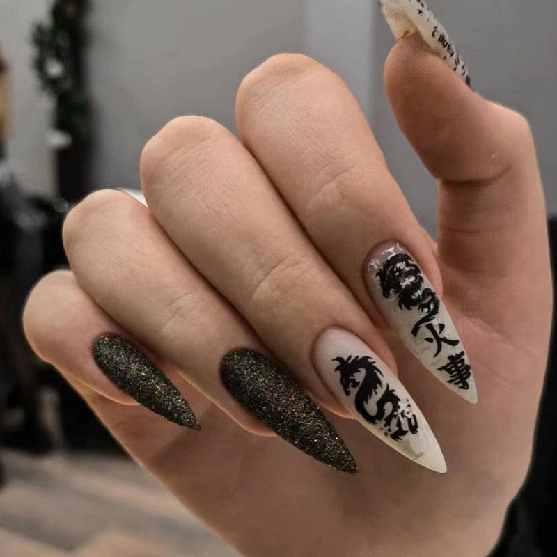 Pointed press-on nails mixing dark glitter tips with black dragon artwork and decorative characters on beige bases. The hand is shown against gray fabric.