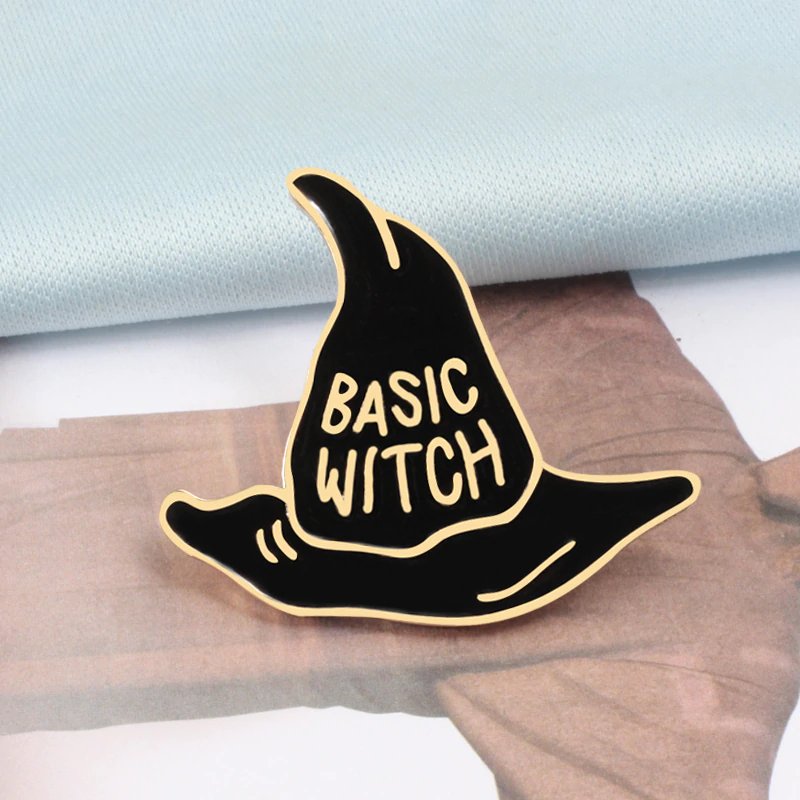 Gold-tone Basic Witch hat pin displayed on printed paper, with bold lettering against black enamel.